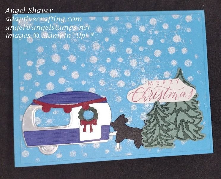 Blue card with heat embossed white dots representing falling snow and a die cut scene with  camping trailer decorated for Christmas with Christmas trees and a black dog