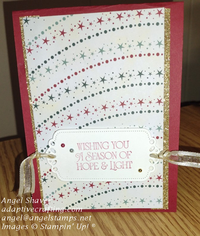 Red Christmas card with patterned paper on front featuring red and green strings of lights and stars backed by gold glimmer paper.  Bottom of card front has sentiment label with gold shimmer ribbon lace through and red, green, and gold pearls.  Sentiment says, "Wishing you a season of hope & light."