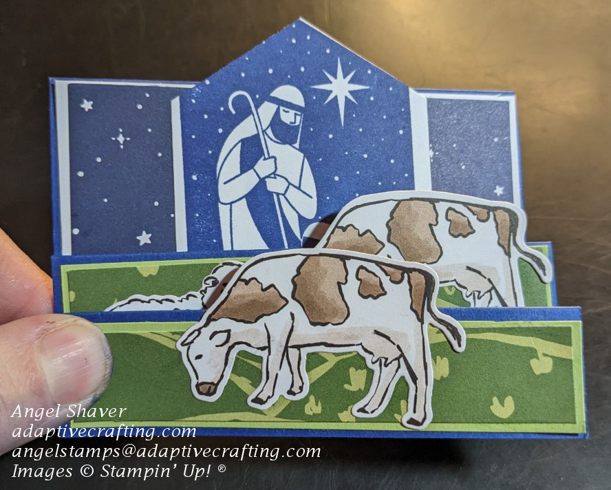 Blue fun fold step card closed.  Largest back panel has stars background paper with stamped image of holy family.  First two steps have rolling hills patterned paper and sheep and cow dies.