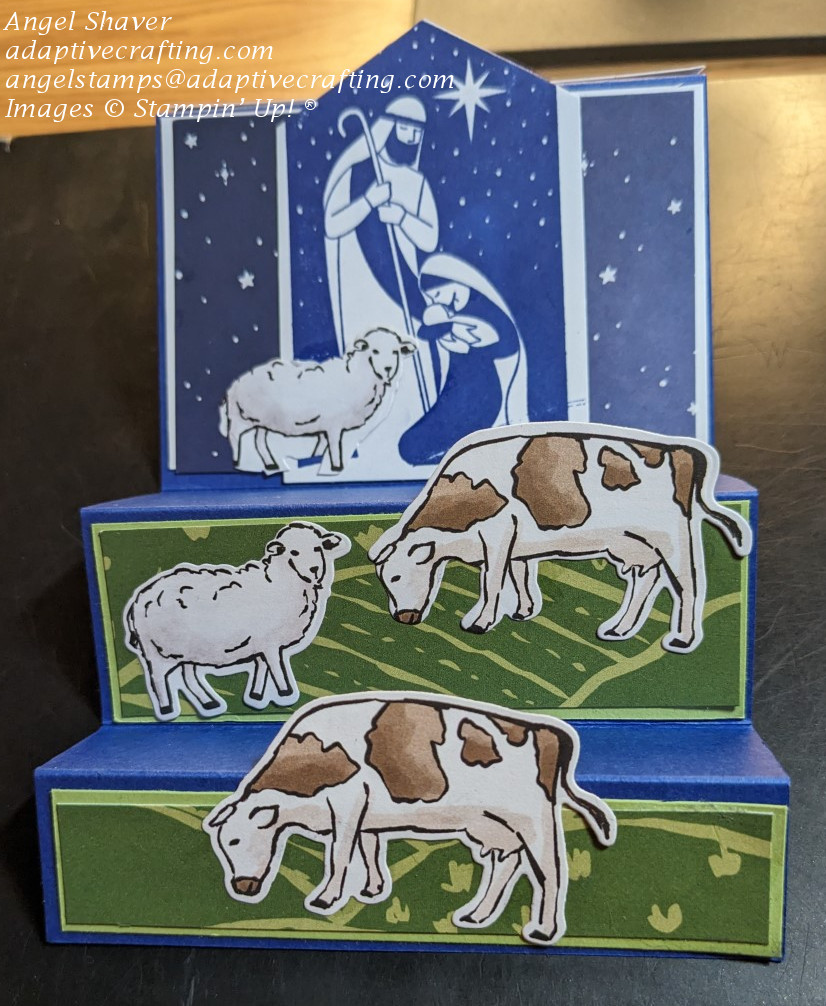Blue fun fold step card with stars background paper with stamped image of holy family with sheep die.  First two steps have rolling hills patterned paper and sheep and cow dies.