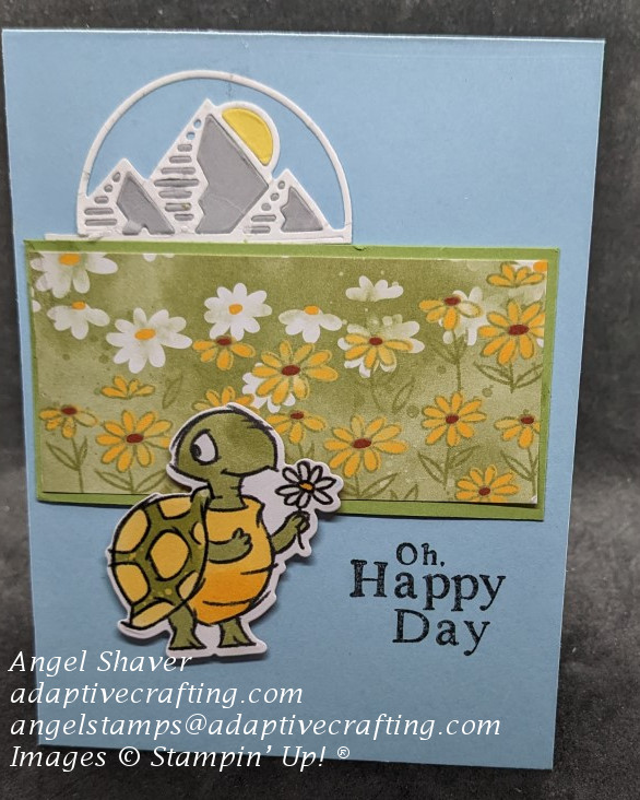 Blue card with strip of patterned paper featuring a field of daisies.  Die of mountain and sun at the top of the card.  Die of turtle holding daisy at bottom of paper strip.  Sentiment says, "Oh happy day"