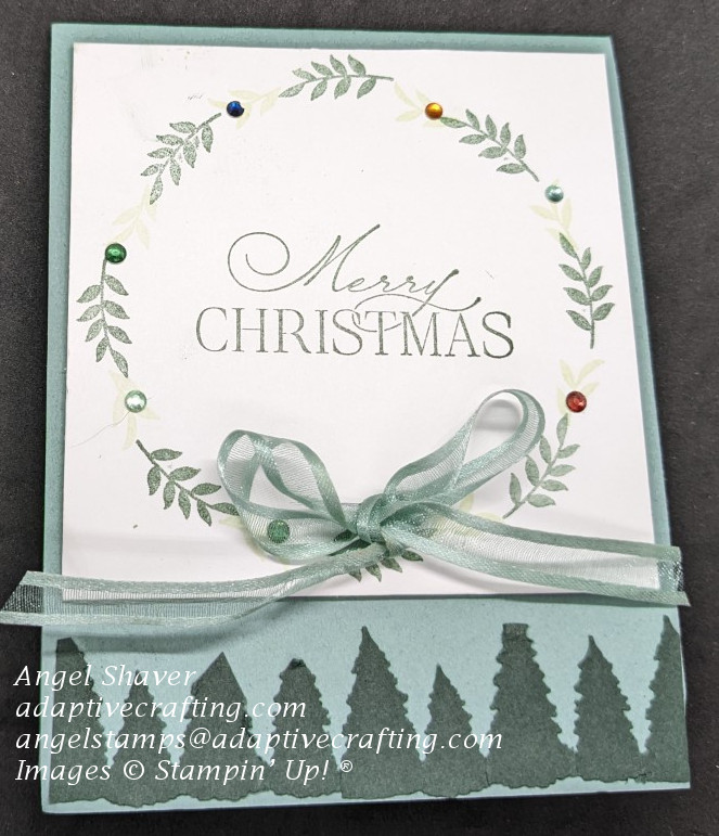 Green Christmas card featuring a wreath made of willow branches, decorated with rhinestones and ribbon.  Dark green evergreen trees make border at bottom of card.  Sentiment says, "Merry Christmas" in the center of the wreath.
