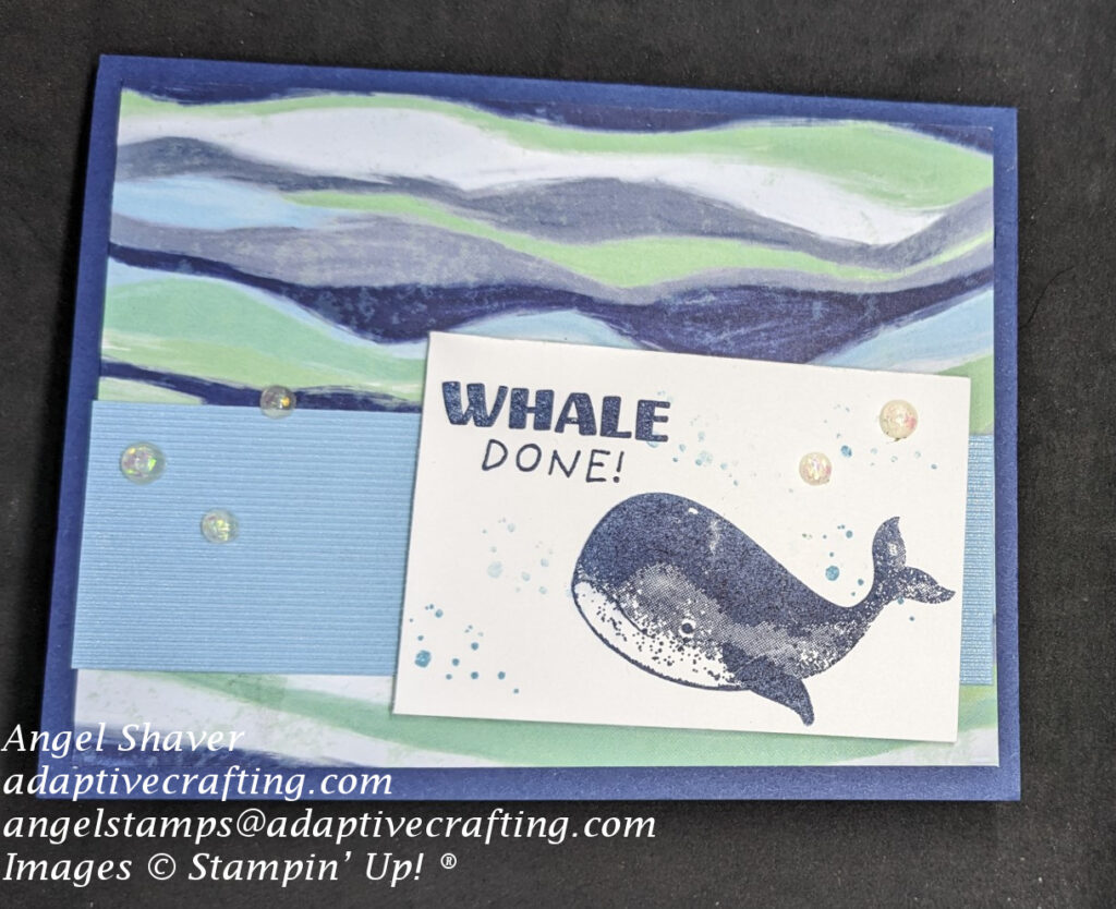 Blue card with patterned paper in blues and greens that looks like waves.  Strip of textured blue paper at bottom of card front.  Top rectangle has stamped whale with bubbles and sentiment that says "Whale Done."  Opal rounds are added to card front.