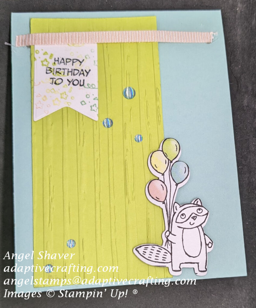Blue card with green embossed paper with raised stripes.  Pink ribbon across top of card with banner hanging down stamped in pastel colored confetti and sentiment says, "Happy Birthday to You."  Die cut of raccoon holding pastel colored balloons at bottom right of card.