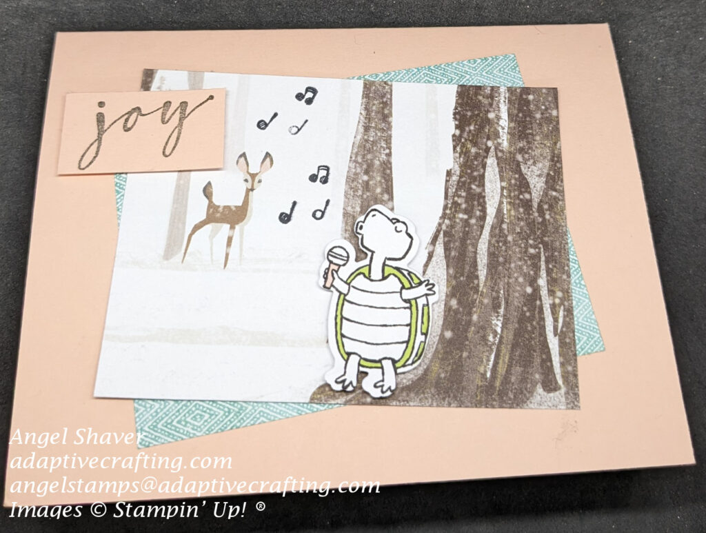 Pink card with two rectangles of patterned paper on card.  A green patterned one is angled behind one with a snowy woodland scene with a fawn.  Music notes are stamped in the woods and there is a die cut turtle singing with a microphone.  Sentiment label says "joy."