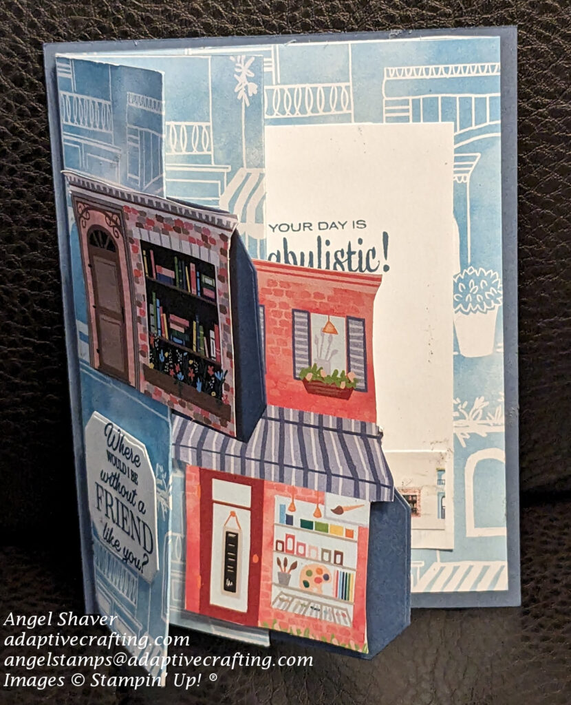 Fun fold 2,4,6,8 blue card featuring Les Shoppes blue patterned paper with white store outlines. The card highlights  a book store and an art store in full color on tabs of card.  Card is blue