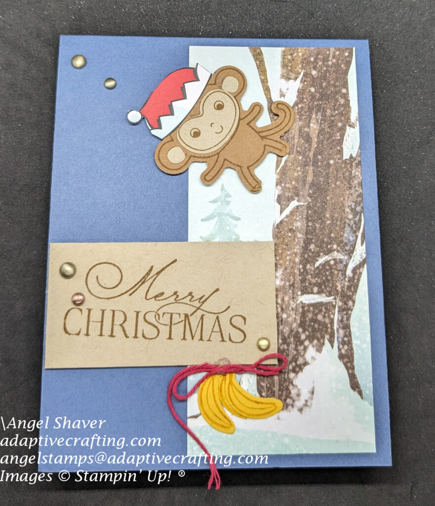Blue card with strip of patterned paper featuring a large tree trunk in the front and evergreen trees behind.  A punch art monkey wearing an elf hat is swinging from a branch in the tree.  There is a bunch of bananas tied with a bow at the bottom of the tree.  Sentiment label says, "Merry Christmas."