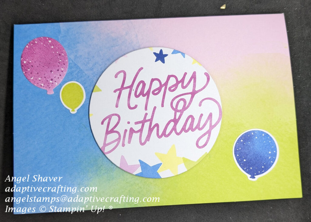 Bright birthday card with splashes or blue, pink, and yellow on card background.  Circle die cut with stars that says, "Happy Birthday" in card center.  Balloon stickers decorate front.