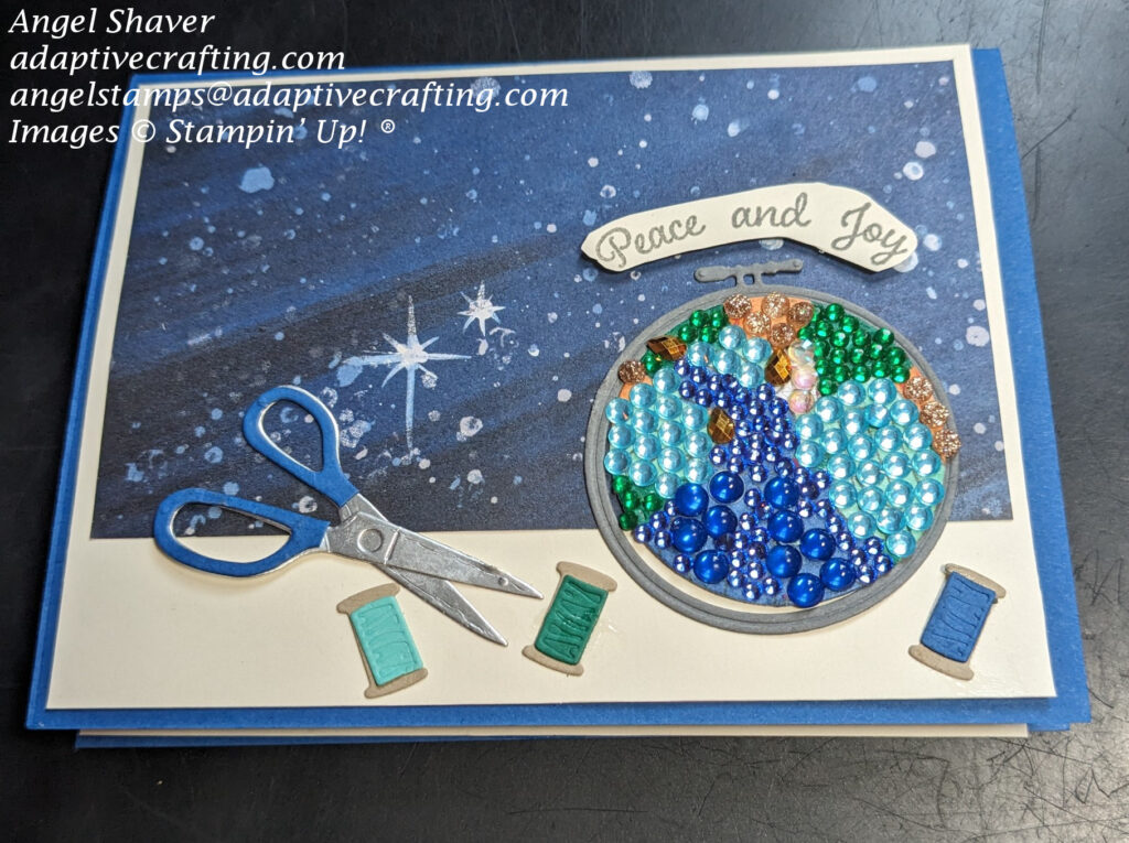 Blue Christmas card with starry sky background paper;  Embroidery hoop with stamped stained glass angel embellished with leftover diamond painting beads and Stampin' Up! embellishments.  Card decorated with scissors and spools of thread.  Sentiment says, "peace and joy."