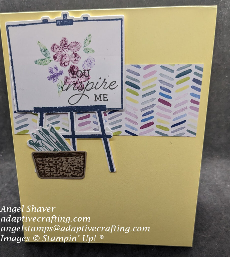Yellow card with strip of patterned paper with angled dashes om a variety of colors.  The focal point image is a diecut easel with what looks like watercolored flowers and says, "You inspire me."  At the bottom of the easel is a basket filled with painbrushes.