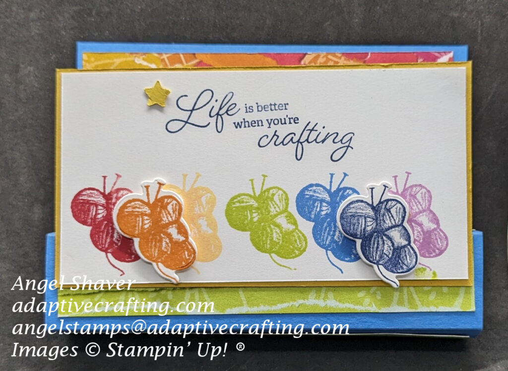 Blue pop up card with patterned paper strips that look like torn paper in rainbow colors covering back layer of card front and the sides of the pop up rectangle.  The featured image on the pop out banner is seven bundles of yarn stamped in the seven different colors of the rainbow--ROYGBIV.  Sentiment says, "Life is better when you're crafting."