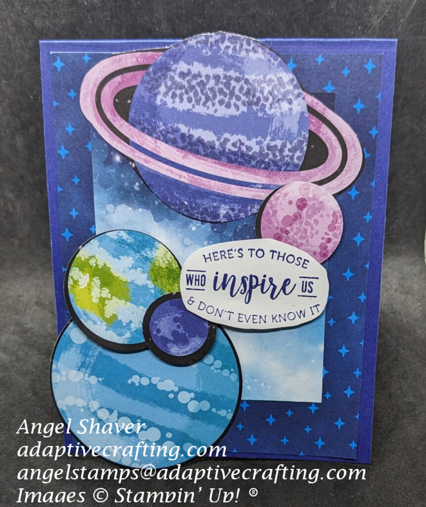 Blue card with two layers of starry background paper.  Five planets are cut out and layered on top of card front.  Sentiment oval says, "Here's to those who inspired us and don't even know it."