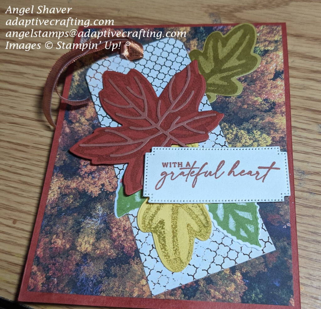 Cajun Craze card with background photo of forest of trees with colored autumn leaves.  Rectangle tag is on top of background layer and has diecut leaves on tag with copper ribbon through top.  Sentiment label says, "with a grateful heart."