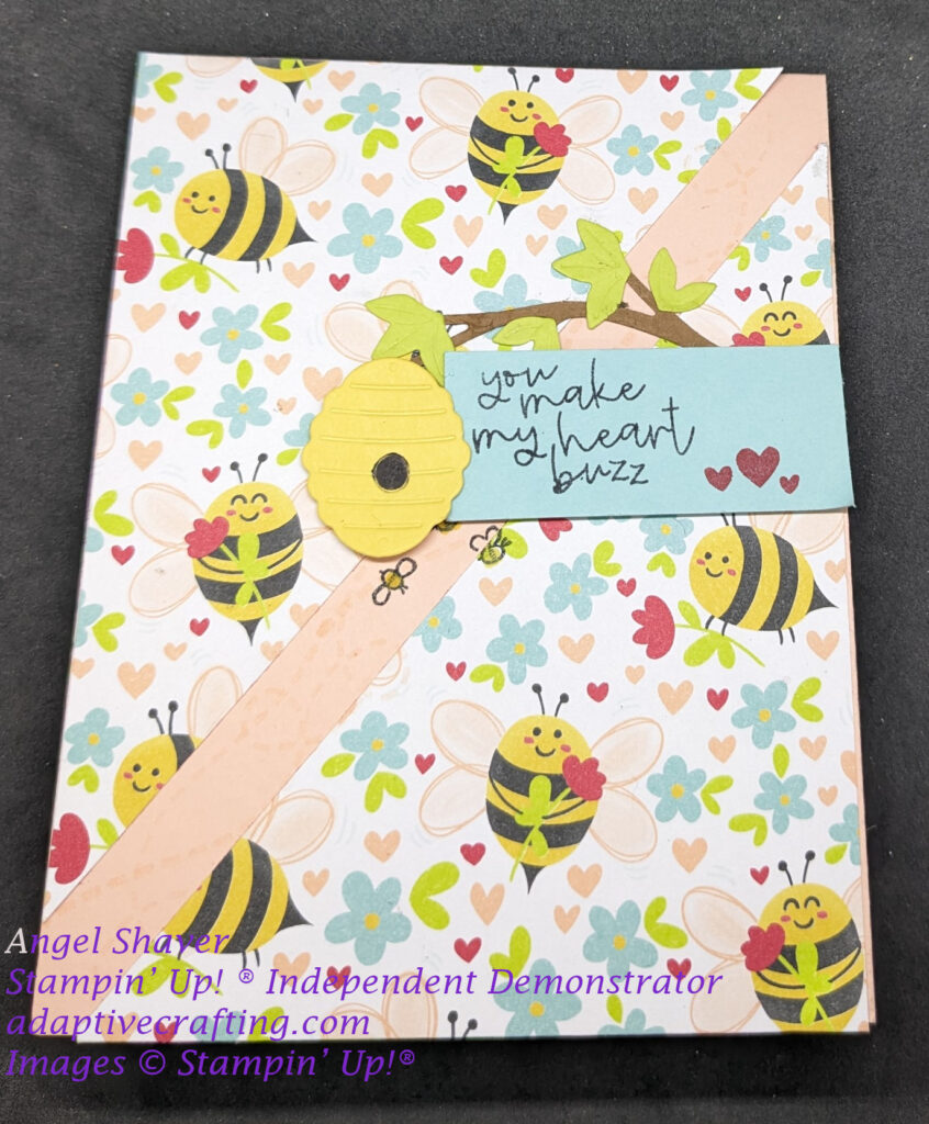 Card featuring two triangle of patterned paper with bees, hearts, and flowers .  Stripe of pink card base can be seen going up the middle of the card diagonally between them.  It is stamped with pink flight pattern lines.  A blue rectangle sentiment label says, "You make my heart buzz." and has three small red stamped hearts.  There is a diecut branch with green leaves and yellow bee hive hanging off sentiment label.  Smaller bees are stamped flying around the bee hive.