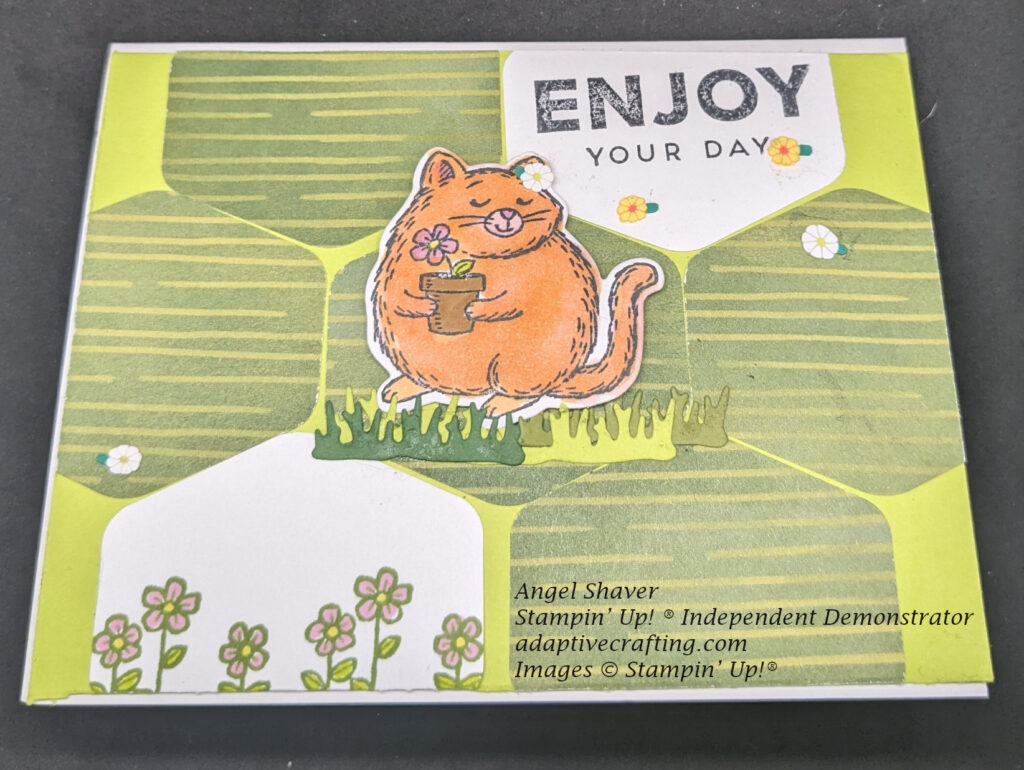 Lime green card front with green striped hexagon punches creating the background.  There are two white hexagons: one with stamped flowers, one with a stamped sentiment "enjoy your day."  Center hexagon has fluffy cat die cut standing on grass dies.  Card front is decorated with white and yellow daisies.