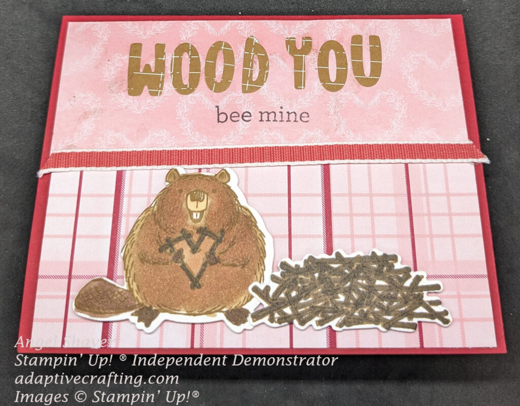 Red Valentine card with pink patterned paper on card front--pink with white hearts on top half of card and pink and red plaid on bottom.  Top half of card featured letter dies that spell out, "Wood You" with a stamped "bee mine" underneath.  Die cut beaver holding heart made of sticks next to pile of sticks on bottom half of page.  Sweet Sorbet ribbon runs across middle of page.