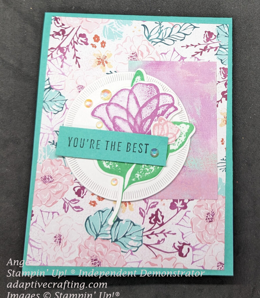 Turquoise card featuring a front panel of patterned paper with flowers and leaves in all five 2024-2026 In Colors.  There is a rectangle with color splashes of purple, blue, and pink.  The top focal image is a die cut circle with diecut translucent purple and pink flowers with green leaves.  The sentiment stamped on a turquoise rectangle says, "You're the best" and there are several iridescent rhinestones on the focal  point die cut circle.