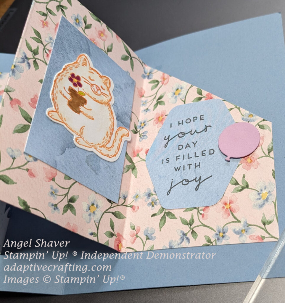 Inside of blue card with w fold insert created with pink floral patterned paper.  Close up of right two sections of the inside.  The first section has a blue rectangle which frames a cat holding a flower pot.  The final section is a pocket for a gift card and is decorated with a blue hexagon that says, "I hope your day is filled with joy" and has a purple balloon die.