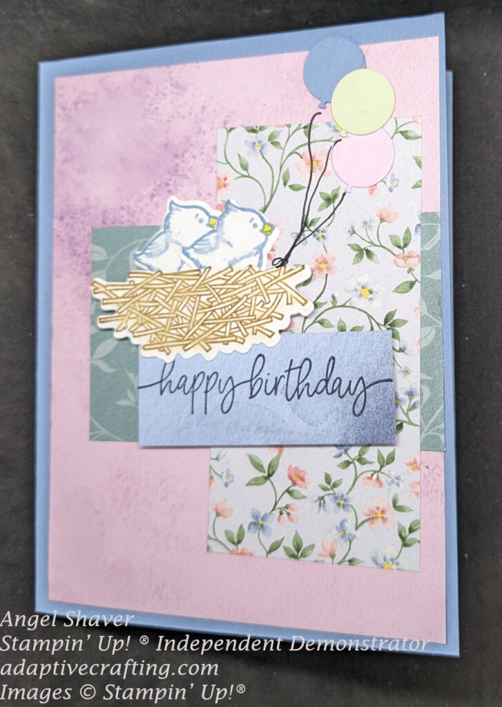 Blue card with all soft pastel colors.  Purple background layer with layers of patterned paper with green leaves, and pink and blue flowers.  Featured images are two diecut birds in a nest with three diecut balloons.
Sentiment says, "happy birthday."