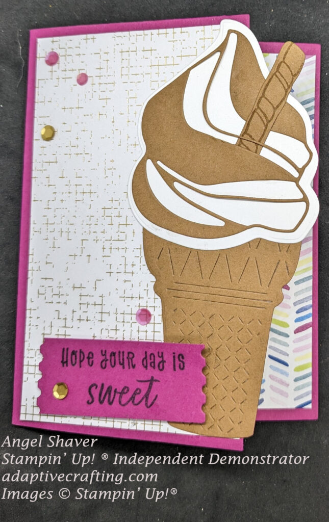 Pink card with gold and white plaid paper on front of card with chocolate and vanilla swirl ice cream cone diecut on the right edge.  Patterned paper with bright, multi colored dashes from inside of card shows behind cone diecut.  Sentiment label says, "Hope your day is sweet."  Card is embellished with pink and gold sequins.