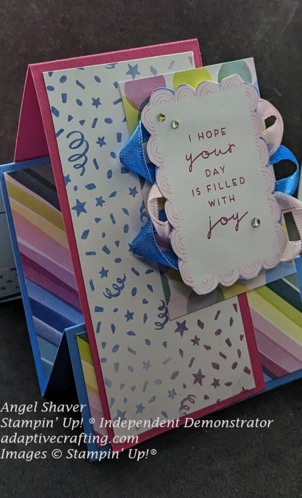 Fun fold faux step card with blue steps covered in diagonal rainbow patterned paper.  Tent is dark pink with confetti patterned paper.  The front focal point is a rectangle with colored circles covered my white rectangle with pink scallop frame.  Blue and pink ribbon loops are between them.  The sentiment says, "I hope your day is filled with you."  Card decorated with rhinestones.