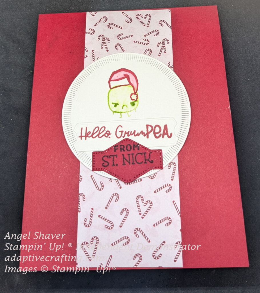 Red Christmas Card with strip of patterned paper with candy canes up the middle.  Then, a white circle with small dashes around outside edge is in center of card.  Green frowning pea is stamped in the circle die with a Santa Claus hat stamped on top.  White sentiment label  says, "Hello, GrumPEA" in red.  A red label underneath says, "From St. Nick."