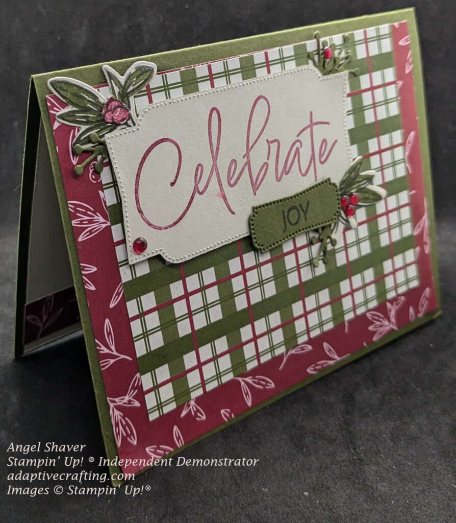 Green Christmas card with layer to red patterned paper with white leaves.  Then red, green, and beige plaid patterned paper.  On top, there is a beige label that says "Celebrate" in red with a green label that says, "Joy".  Around the label are green leaf dies and red berries.  