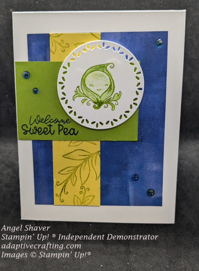 White baby card with blue background paper, with a yellow strip of paper stamped with green pea leaves running vertically on blue paper. Green rectangle lays on top horizontally with sentiment "Welcome Sweet Pea".  A white circle die with a decorative cut edge and stamped single pea in pod is the top focal point image.  Card also has five blue and green tinsel dots  