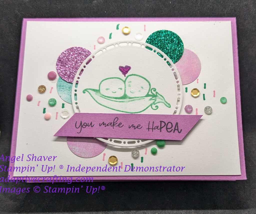 Purple card with white layer will lots of circle 1" punches, sequins, and other circular embellishments in pink, purple, green, gold, and white.  White circle die with cut out frame around circle is stamped with two peas in a pod with a heart between them.  Purple sentiment label on top says, "You make me HaPEA."