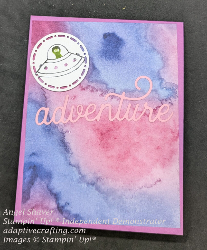 Pink card with patterned paper with pink and purple splotches.  A pink die cut word "adventure" is across the middle of the page with a white die cut circle stamped with an alien in a spaceship about it.