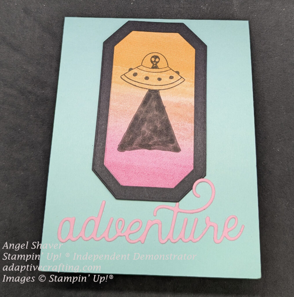 Light blue/green card with pink word die "adventure" at the bottom of the card front.  A Black frame is above that with patterned paper inside the frame with and orange changing to pink background.  An alien in a spaceship is stamped in black with a solid black ray beam coming from the spaceship