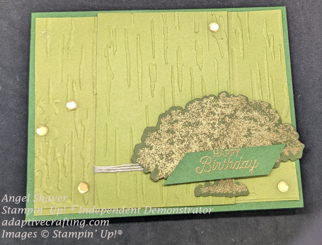 Birthday card in shades of green.  Card front layer embossed to look like bark with second panel embossed and raised with dimensionals in the center.  Twine wrapped around center panel.  Die cut tree stamped and heat embossed in gold is at bottom left of card with green sentiment label heat embossed in gold "Happy Birthday::  Gold sequins are added to card front.