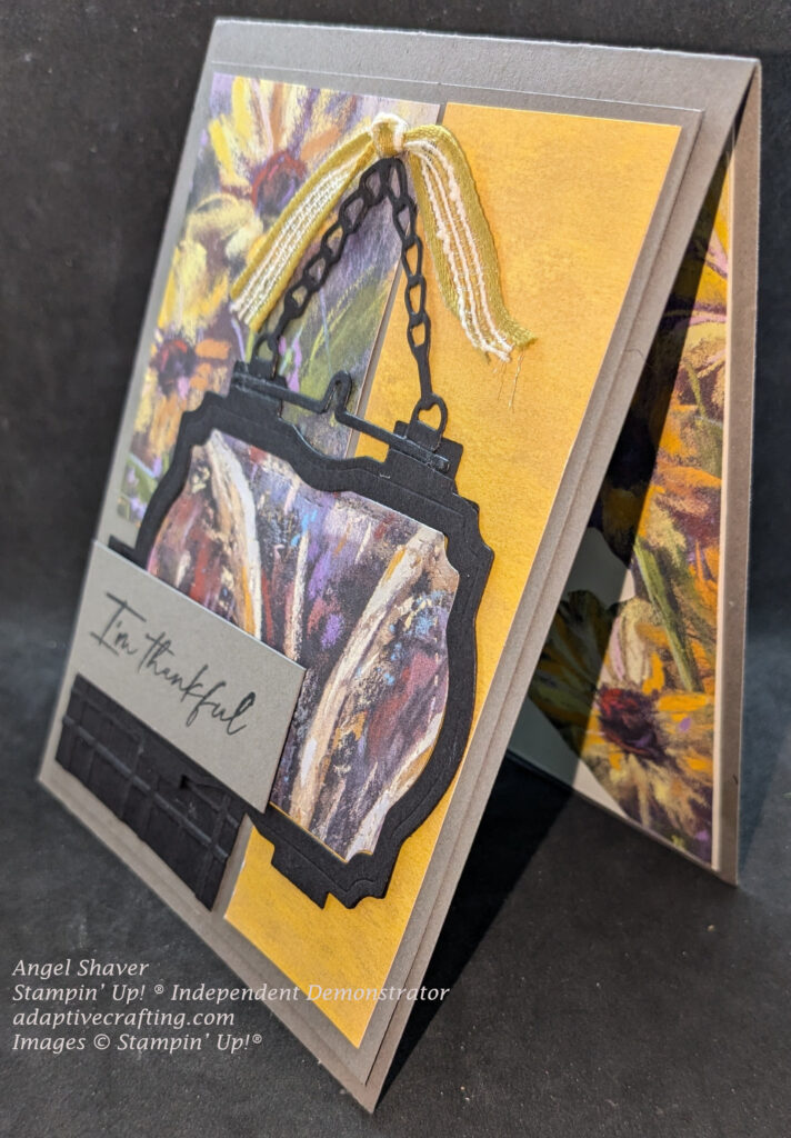 Grey thank you card featuring panels of yellow, black, and sunflowers on the card front.  Featured black frame die with hanging chain frames patterned paper featuring shocks of wheat.  The frame is hanging from a yellow knotted ribbon.  The gray sentiment label says, "I'm thankful."  Card is shown at an angle so you can see sunflowers on inside of card.