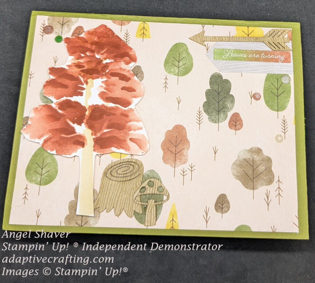 Green card with layer of patterned paper with various colored trees.  There is a tree with red leaves created with layering masks.  Beneath the tree is a wood log die and mushroom.  The sentiment sticker is mutli-colored on a wood patterned label and says, "Leaves are turning."  Wood arrow embellishment is above sentiment.