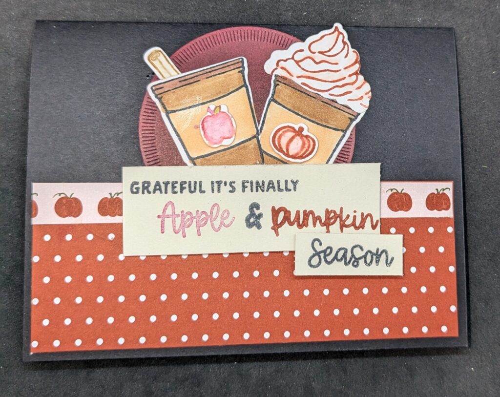 Black card with orange patterned paper with white dots across bottom of card with strip of patterned paper featuring pumpkins on top of that.  In center of top half of card is red circle.  There are two different cups on the circle one with an apple on the label and a cinnamon stick inside.  One with a pumpkin on the label and topped with whipped cream.  The sentiment label at the bottom of the circle says, "Grateful it's finally apple & pumpkin season."