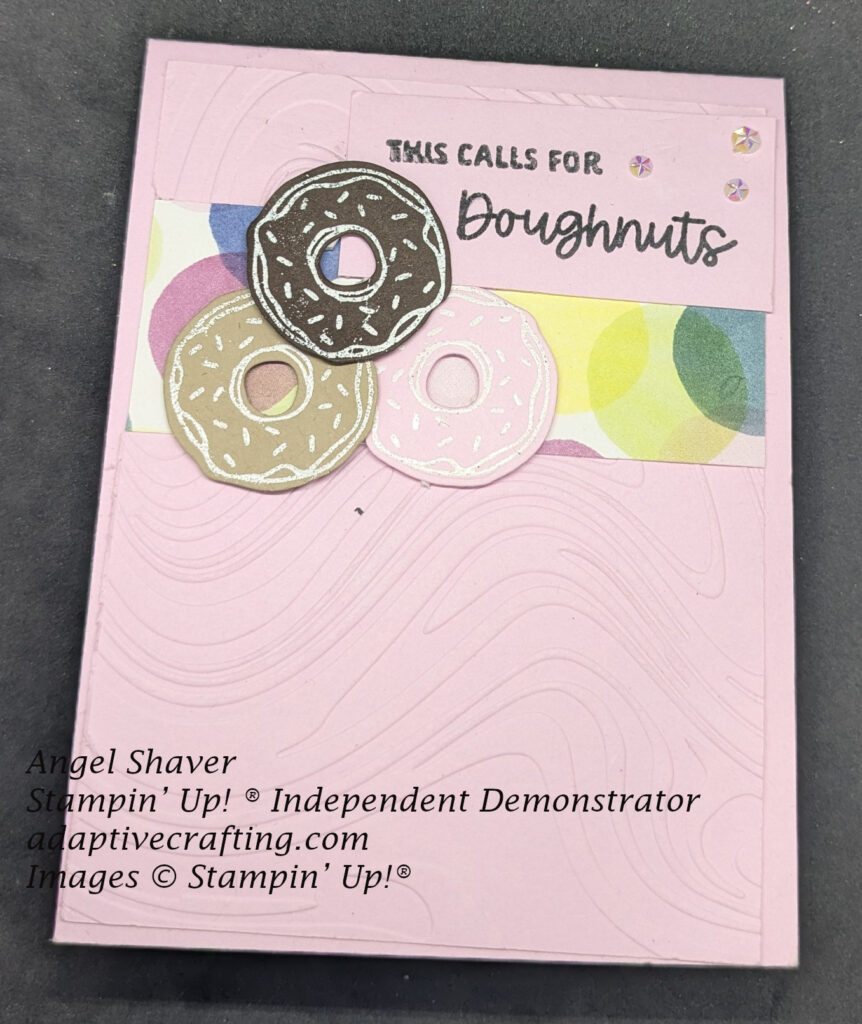 Pink card with layer of pink embossed swirls on card.  Strip of patterned paper towards the top with circles in bright colors--yellow, pink, blue.  There are three donuts heat embossed with white sprinkles.  One dark brown, one light brown, and one pink.  The sentiment label says, "This calls for doughnuts." 