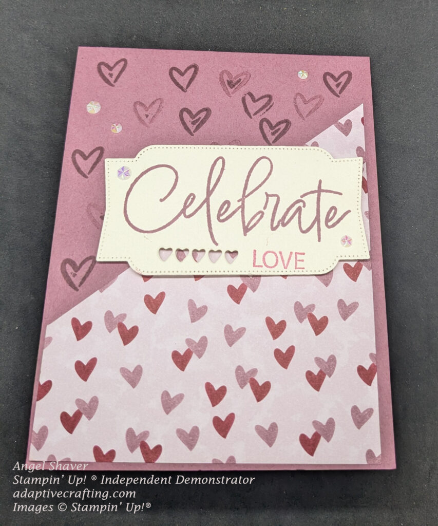 Pink card with lighter pink patterned paper with pink and red hearts cut diagonally and glued to the bottom half of the card.  The top half is stamped tone on tone with heart outlines.  The center of the card holds the focal point element, a cream colored label that says, "Celebrate love."  There are five tiny heart shapes cut from bottom of the label.  The card is decorated with clear faceted gems.