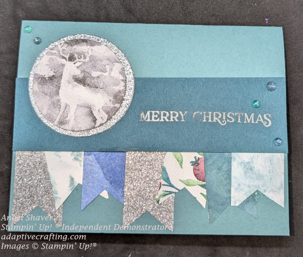 Teal Christmas card with strip of darker teal cardstock across center of card.  That has a silver heat embossed sentiment that says, "Merry Christmas". Flags are hanging from darker cardstock strip n silver glitter, blue and green DSP, and DSP patterns of trees and leaves and berries.  Focal image is a circle of DSP with a deer on it framed with a circle of silver glitter.