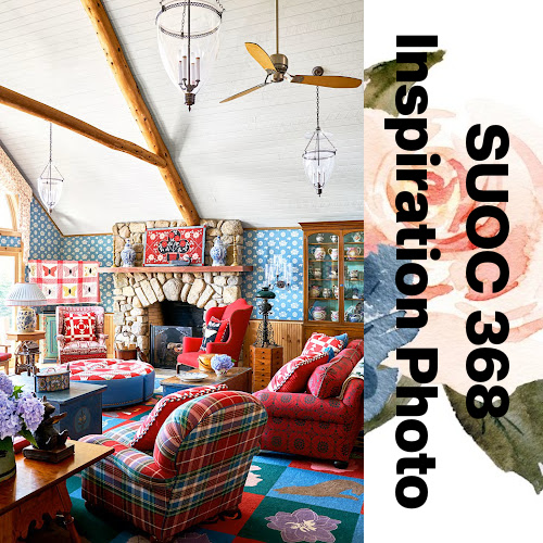 SUOC368 Inspiration Photo.  Picture of living room with stone fireplace, large stuffed red patterned couch, and red and green plaid chair.    The room has wood beams on the ceiling, along with a ceiling fan, and hanging lights.  There is a quilt of butterflies.  A wooden shelf full of vases and other decorations.  There is a vase of purple flowers on one table plus several other tables and a round ottomon in the middle of the room.