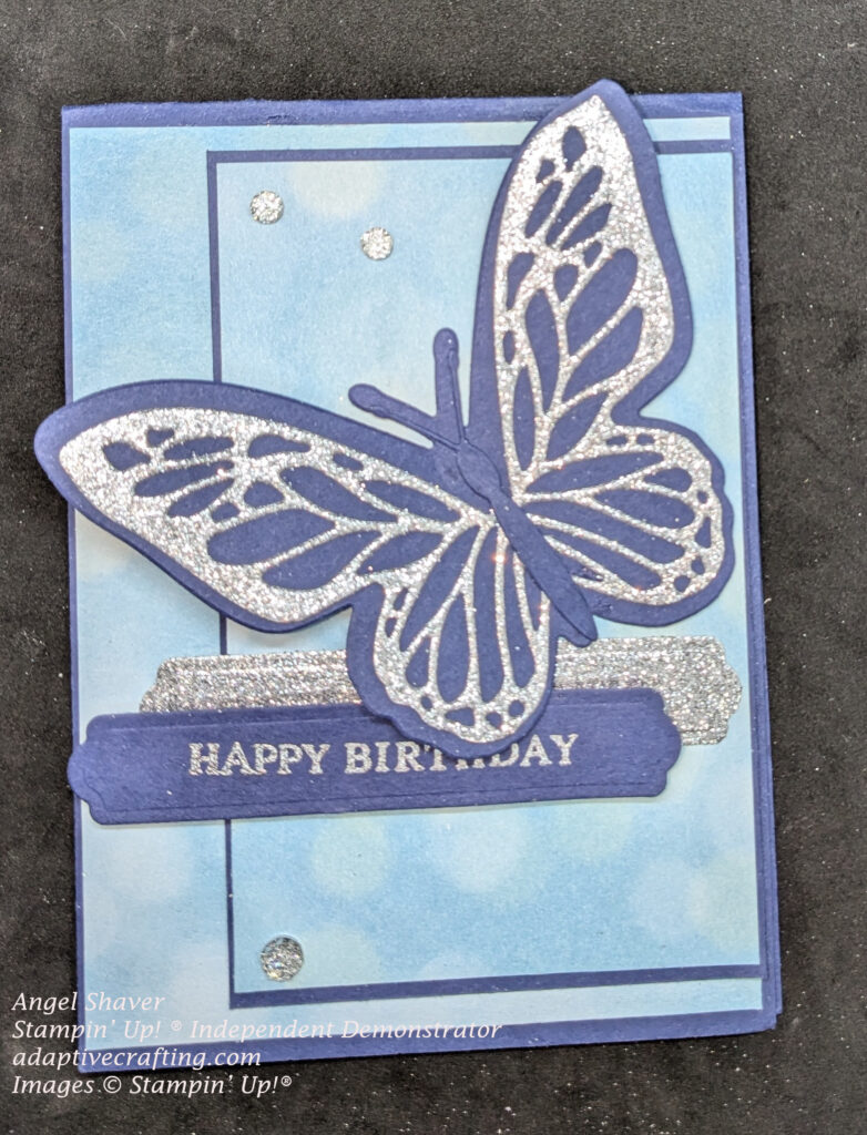 Blue card with light blue bokah dot patterned paper.  There is a large blue butterfly diecut with silver diecut wing details.  A silver glitter label lays behind a blue sentiment label heat embossed in silver to say, "Happy Birthday."  There are silver embellishments finishing off the card.