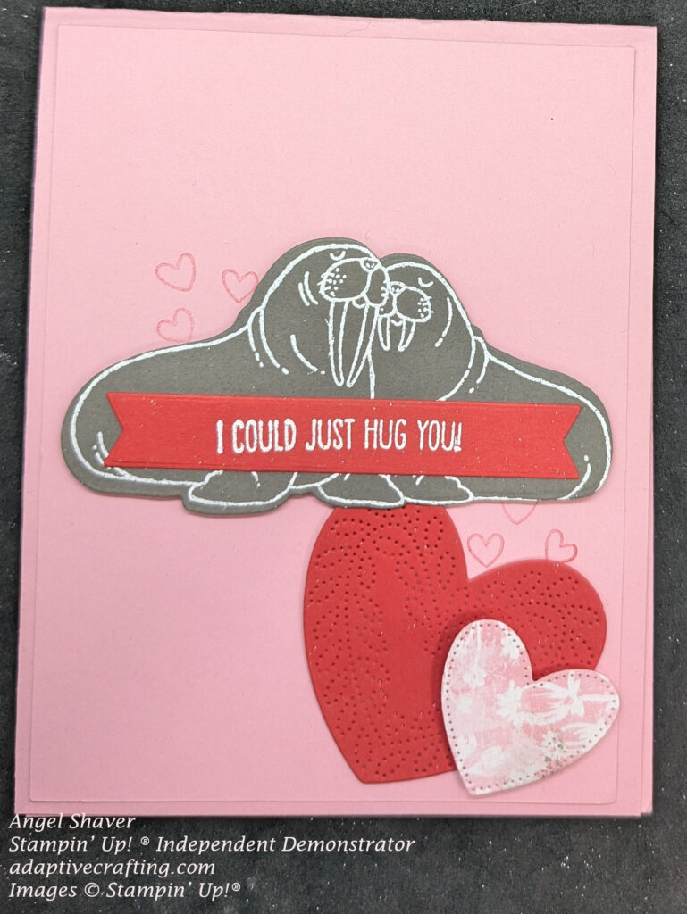 Pink card with red hearts stamped above and below walruses.  Diecut of two walrus is heat embossed in white on gray paper and centered on the card.  Two heart dies are place below the walruses.  One is a red heart with a leaf frame cut into it.  There is a smaller heart cut from pink patterned paper with white flowers.  There is a red sentiment label on top of the walrus that says, "I could just hug you."