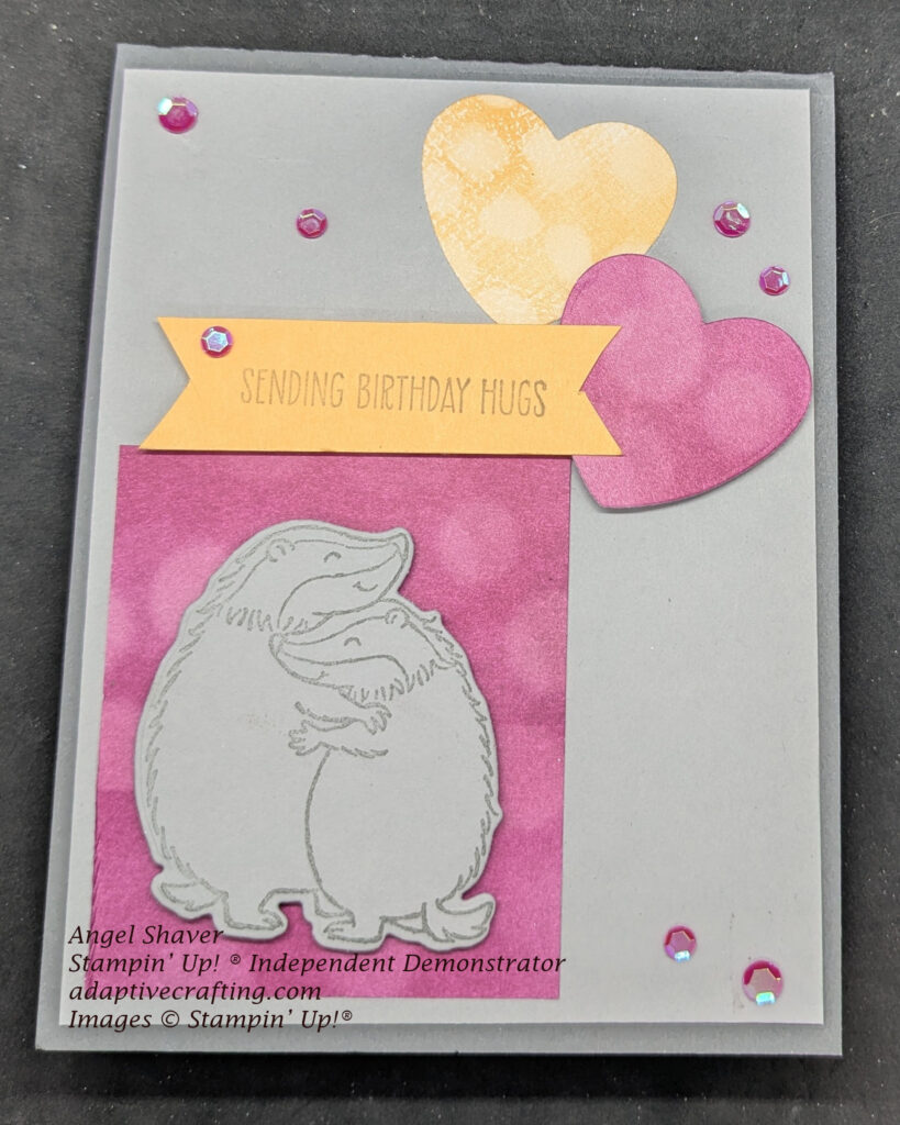 Gray birthday card featuring a stamp of two hugging badgers positioned on a retangle of purplish bokeh paper.  There are two bokeh hearts--one purplish and one peach.  The peach label says, "Sending Birthday Hugs."  Purple sequins decorate the card.