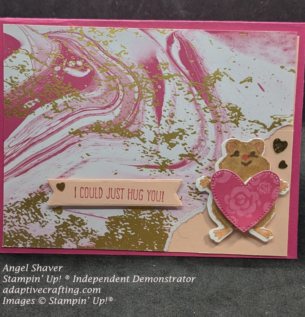 Pink card with swirling card front with pink, white, and gold swirls.  Bottom right corner is torn to reveal lighter pink underneath.  Hamster die holding a pink heart with roses is in the torn corner.  Sentiment label says, "I could just hug you!"  Gold heart sequins embellish the card.