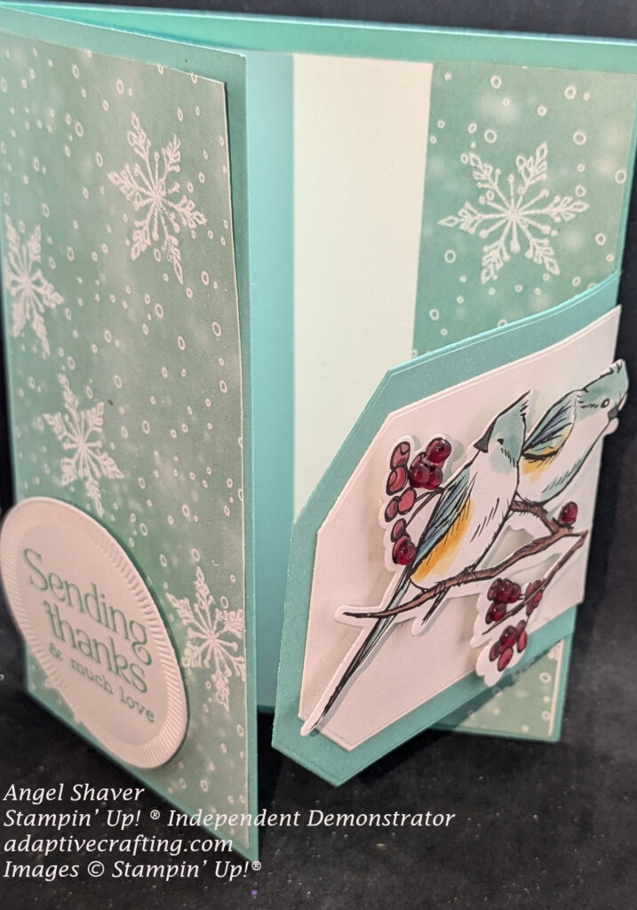 "sending thanks and much love."  Hinge made of countryside corner dies--large one teal with smaller white one on top is hinged to right side of card. Image of two blue and yellow birds on branches with red berries is featured image on hinge, Card is open so you can see inside of card which contains white panel to write on and teal patterned paper strip with white circles and snowflakes on right side of inside panel.