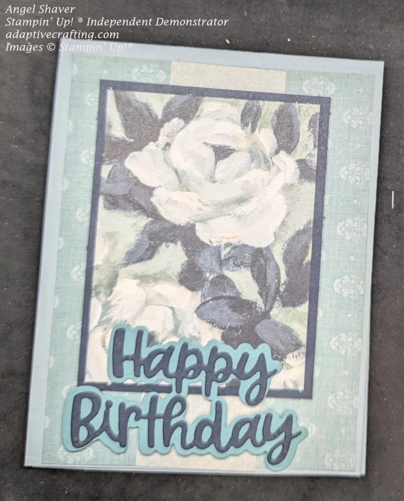 Light blue card with teal patterned paper layer that looks like an antique wallpaper pattern. Strip of light patterned paper down center of layer with a piece of patterned paper that looks like a painting of white roses with dark leaves framed by dark blue paper. The bottom of the card has diecut words that say "Happy Birthday"