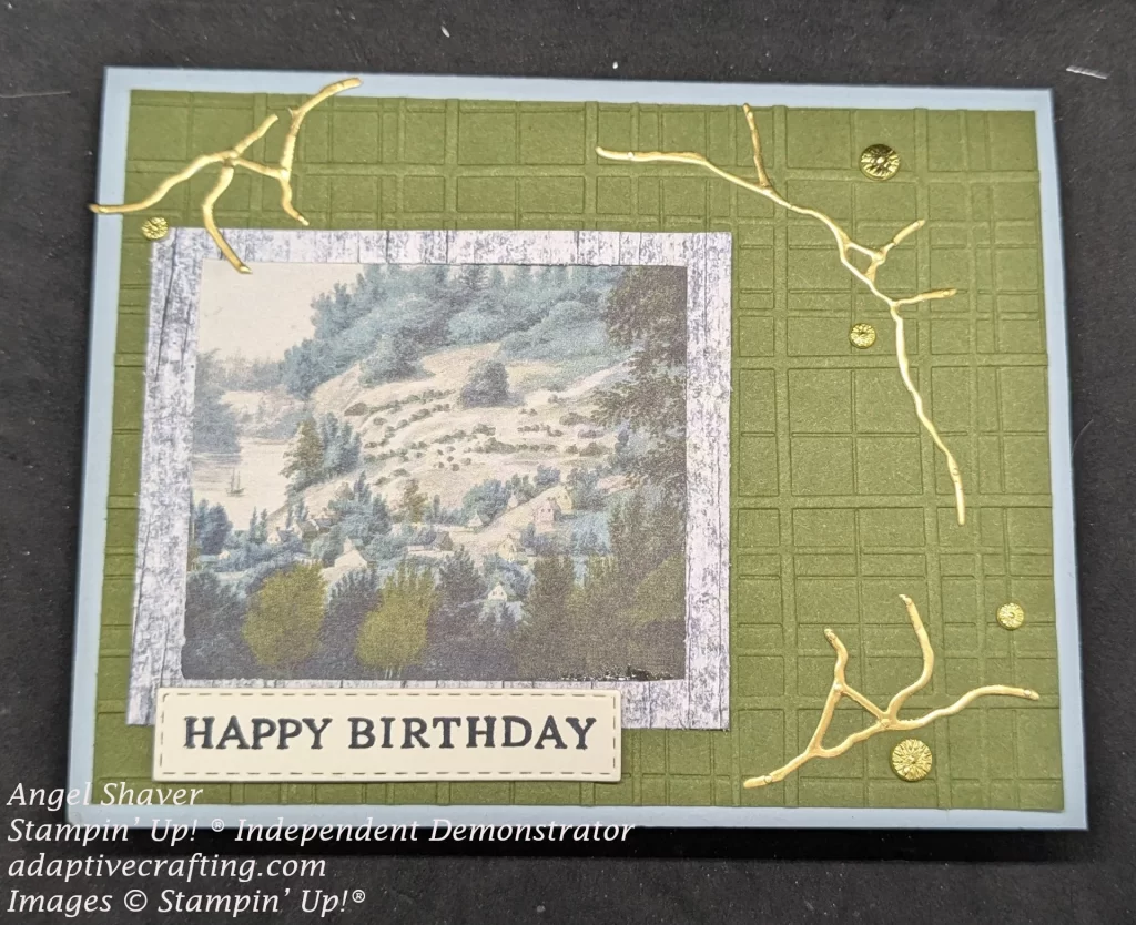 Blue card with green card front layer embossed in plaid. Image of a snowy mountain landscape framed with blue wood frame is the focal point. There are three clusters of gold foil lines with gold floral embellishments. Sentiment label under the framed picture that says, "Happy Birthday"