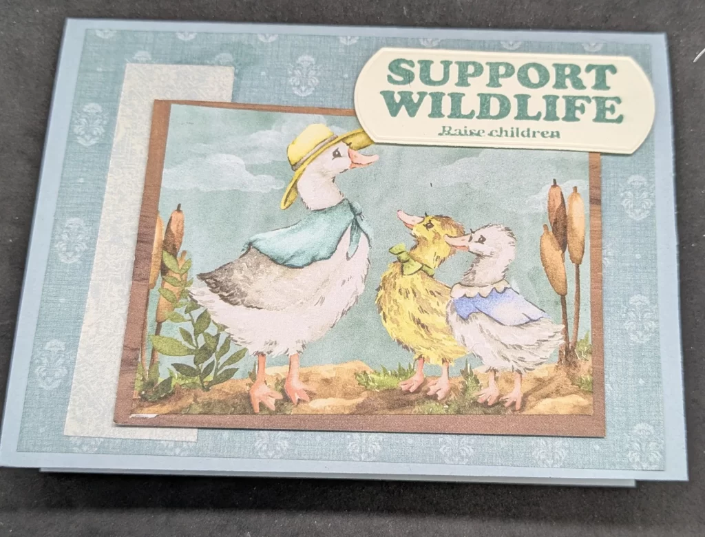Blue card with teal background layer with light blue vintage shapes. Strip of lighter blue patterned paper is on front with focal image layered on that. Focal image is patterend paper scene of Mother goose wearing hat and cape lookin at two gooslings--one yellow wearing a green bow tie and white gosling wearing a blue shawl. They are surrounded by cattails. Photo is framed by brown patterned paper. Sentiment label says, "Support Wildlife. Raise children"
