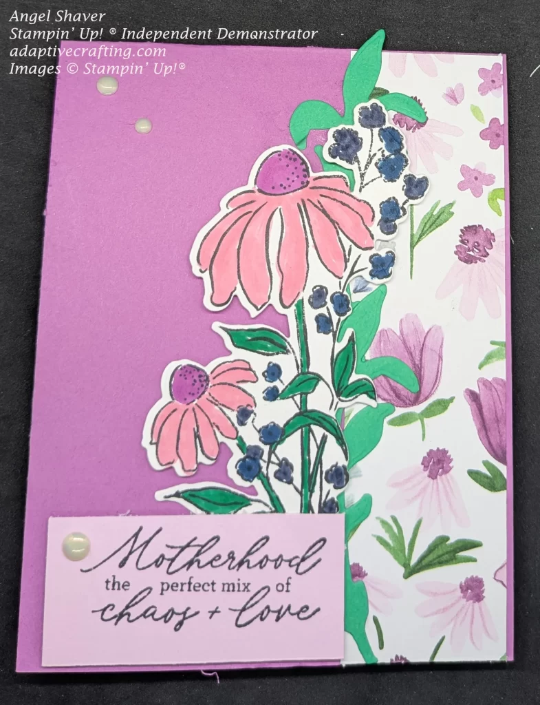 Purple card with strip of paper featuring pink, purple, and blue flowers. Green die cut leaves create border between patterned paper and purple cardstock. Diecut image of pink coneflowers with purple centers and small blue flowers is the focal point of the card. Sentment label says,, "Motherhood is the perfect mix of chaos and love." Card is decorated with vintaage pearls.
