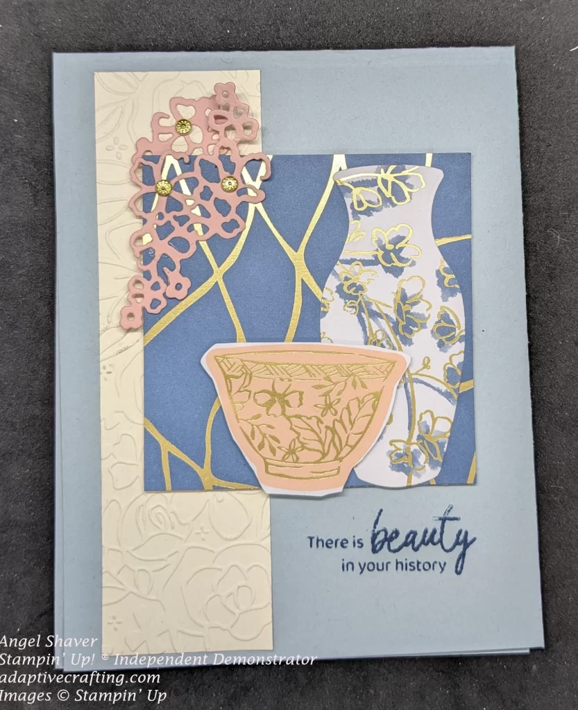 Blue card with cream embossed strip with flowers. The focal image is a blue square with gold streaks. Diecut pink flowers are on the top left corner of the square. There is a tall white vase with blue and gold flowers and a pink bowl with gold flowers. The sentiment is stamped under the patterned paper square and says, "There is beauty in your history."