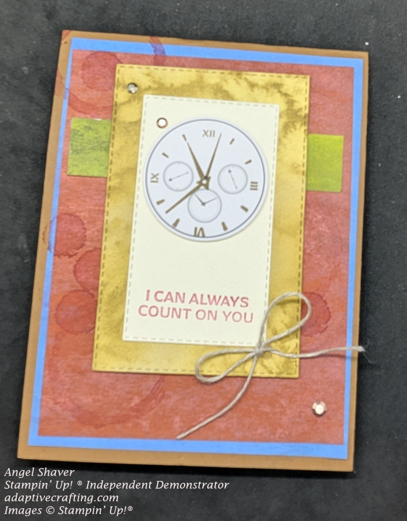 Brown card with blue and red layers. Circles are stamped on red layer. Green strip of patterned paper runs horizontally across card. Stitched rectangle in gold with a cream colored rectangle with "I can always count on you." with clock at top of rectangle. Bow of linen thread is added to bottom of cream colored rectangle. Copper and gold embellishments are added to the front.